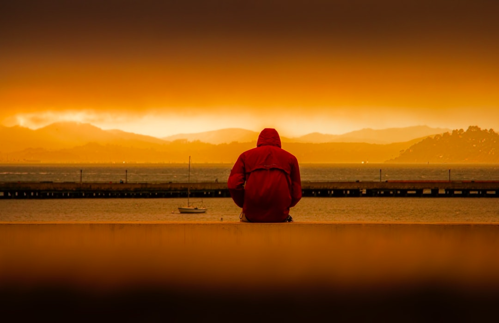 Sad man in red coat sits beside a lake at sunset.