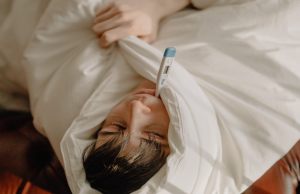 Man in white shirt lying on bed with a thermometer in his mouth