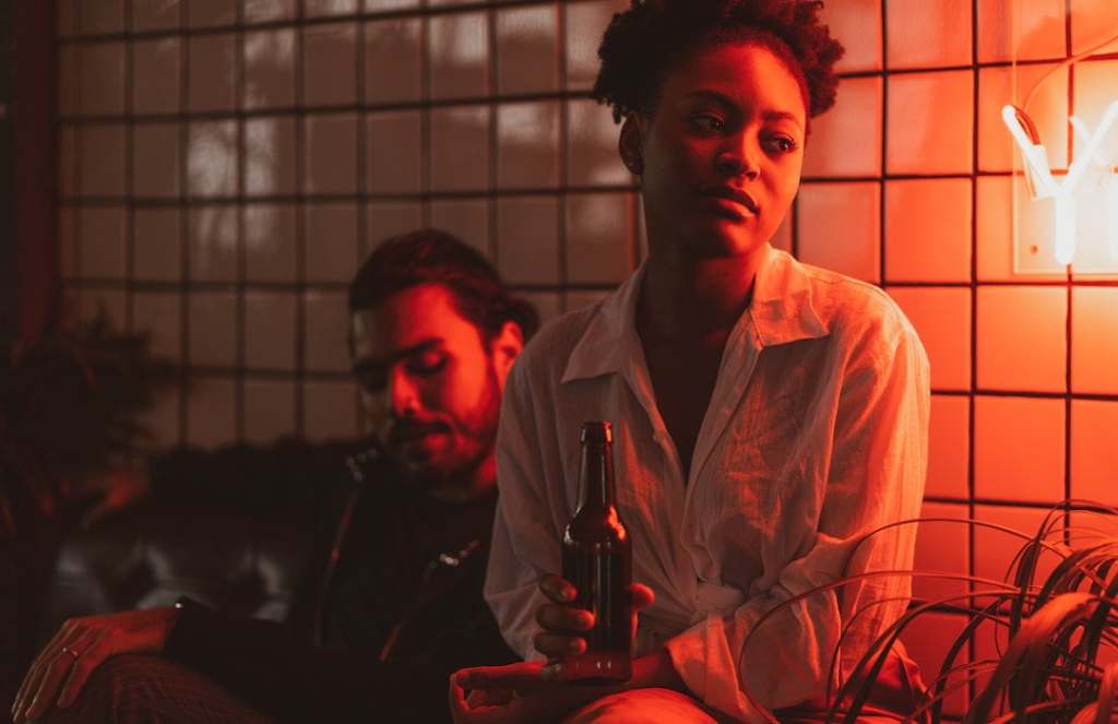 Depressed young woman holds beer in bar with young man in background