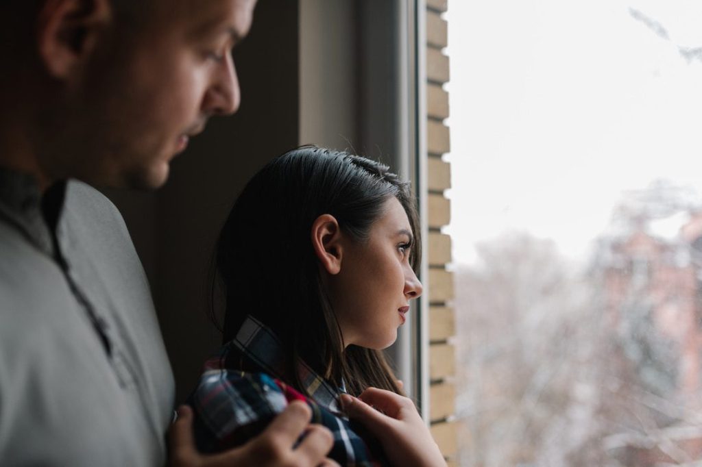 A woman with dark hair looks out of a window on a cloudy day. A man in a gray shirt has his hand on her shoulder, comforting her.