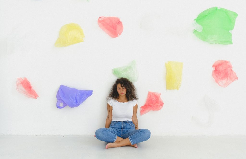 Sad woman sits in white room surrounded by colorful, floating plastic bags