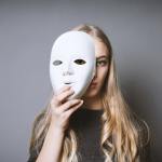 Teen blonde girl hiding her face behind a mask with a gray background