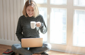 woman in grey shirt sitting on floor with coffee and laptop