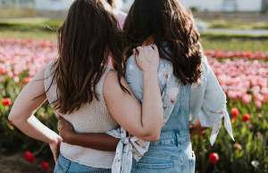 black woman in overalls and green floral shirt hugging white woman in cream knit shirt in garden of pink tulips