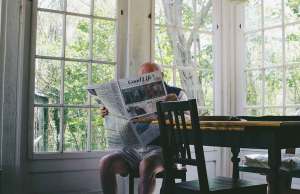 elderly man sitting at wood table holding newspaper by windows