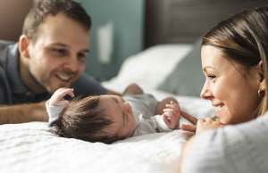 man and woman in grey shirts looking at baby laying on white bed