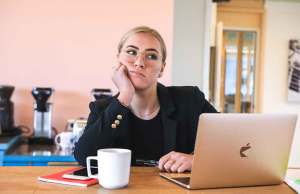 blonde woman in black shirt sitting at desk with Macbook and white coffee mug