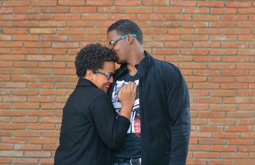 black man and woman wearing black jackets hugging by brick wall