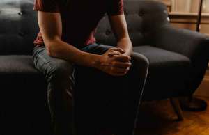man in red t shirt and dark jeans sitting on grey couch holding hands together
