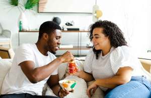 man and woman in white shirts and jeans eating together on couch