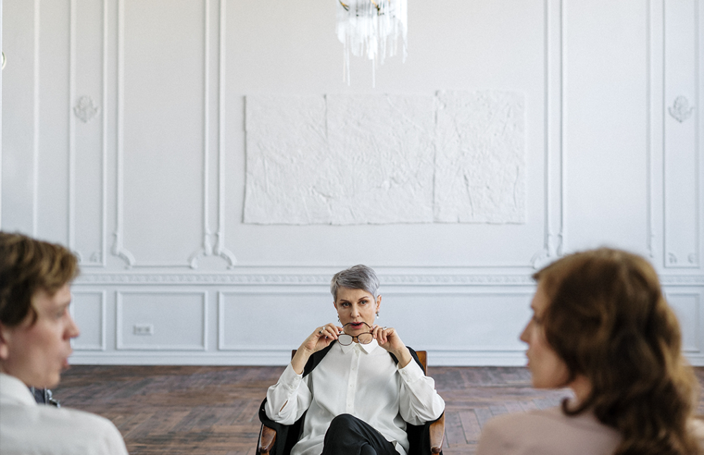 woman in white long sleeve shirt sitting in a chair on wooden floor looking at two individuals