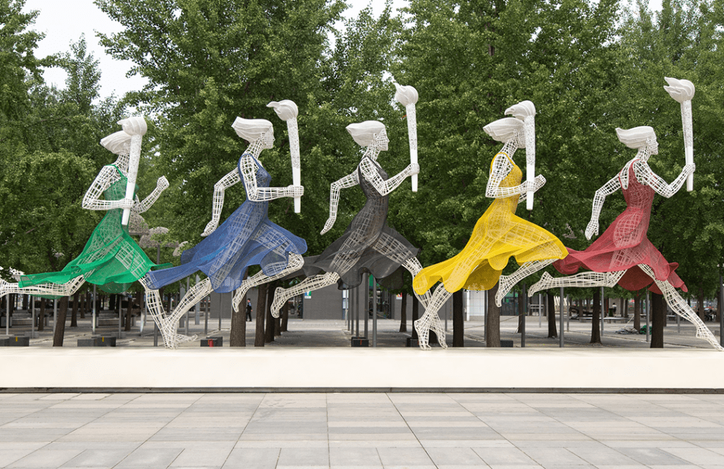 five running women wearing different colored dresses holding Olympic torches