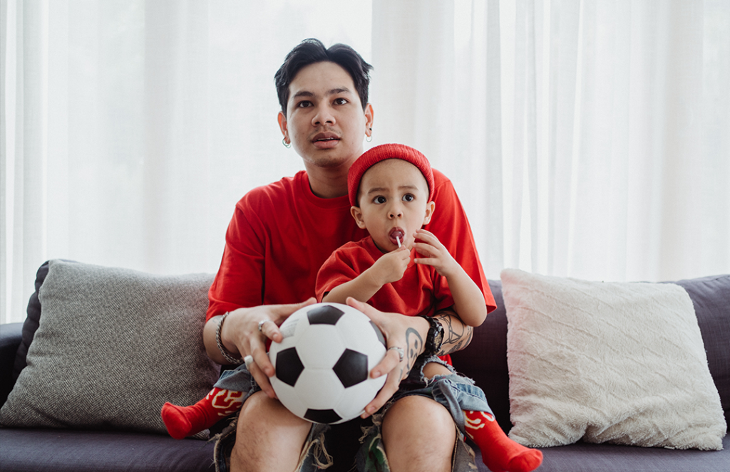 man in red t-shirt sitting on a couch holding a soccer ball and child with a matching shirt