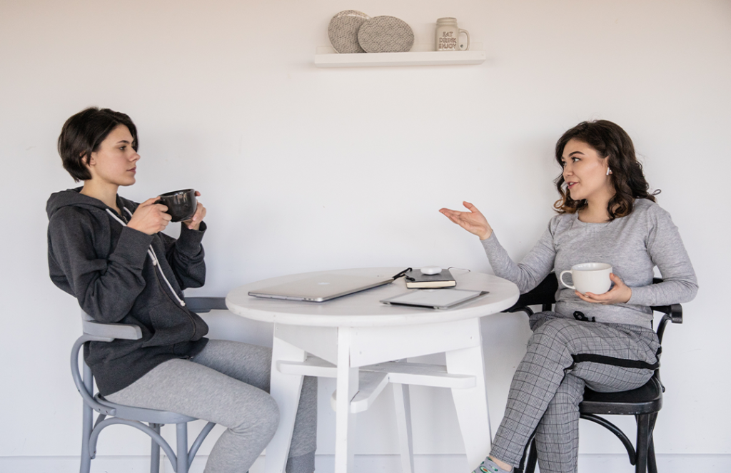 two women sitting at table talking holding coffee cups