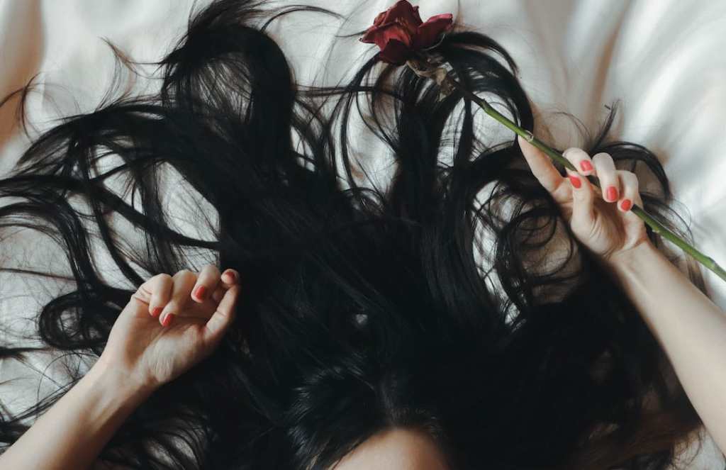 Woman with black hair lies in bed with red rose and red fingernails