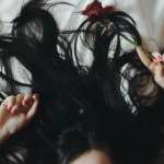 Woman with black hair lies in bed with red rose and red fingernails
