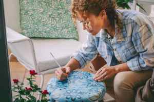 Woman in a blue flannel painting blue designs on a stool