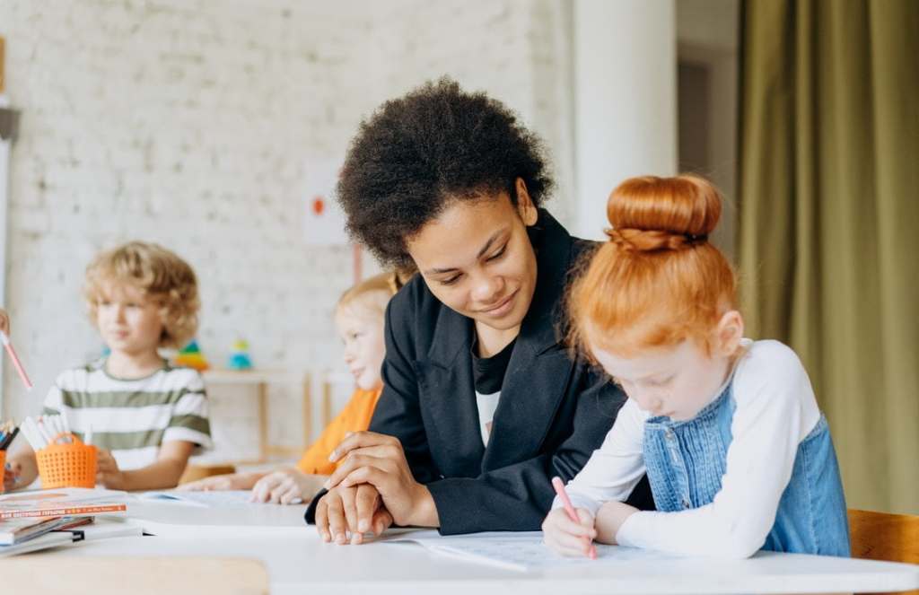 Red-headed girl practices writing as teacher watches in classroom