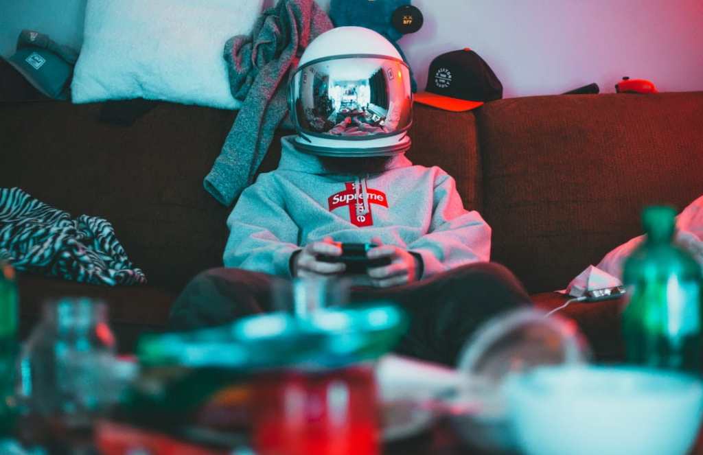 Teenage boy in sweatshirt and helmet plays video games on a messy couch