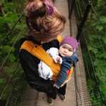 Mom walks baby in baby carrier across suspension bridge in a forest