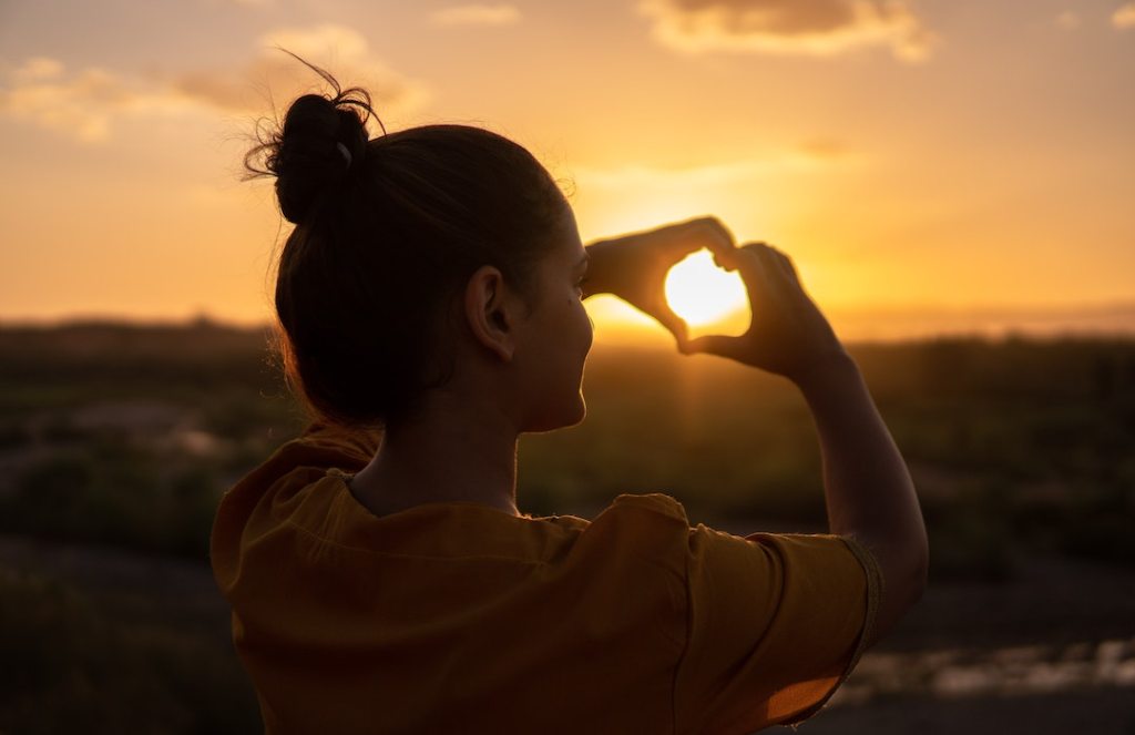Woman in orange t-shirt watching the sunset while holding up a heart with two hands