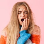 Portrait of a tired young girl wearing sweater standing isolated over pink background, yawning