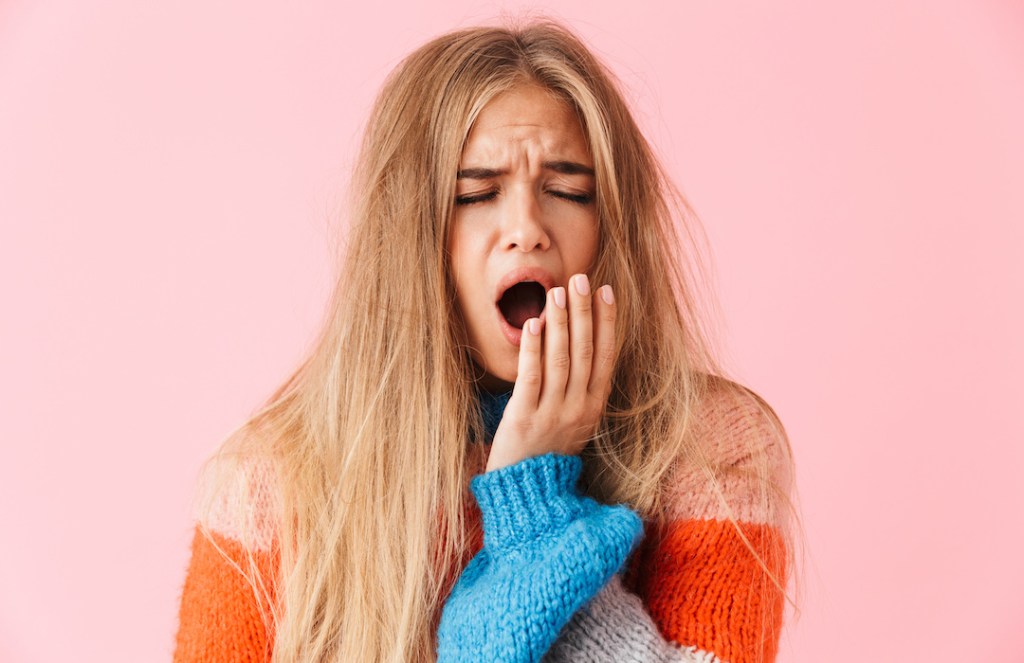 Portrait of a tired young girl wearing sweater standing isolated over pink background, yawning