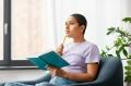 Woman journaling in a blue chair next to a window holding a pencil to her chin