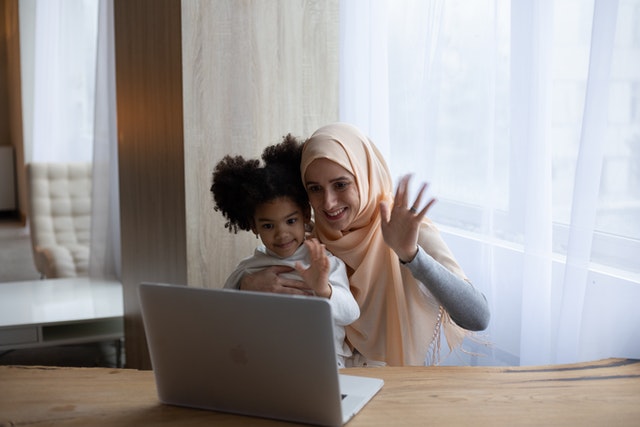 A woman holding her child while talking on a laptop and waving.
