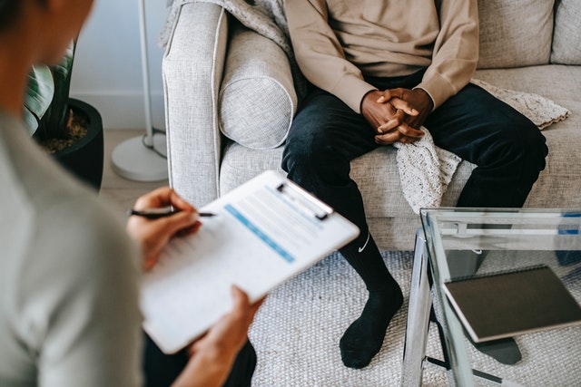 a counselor holding a clipboard and talking to their patient who is sitting on a couch with their hands held together.