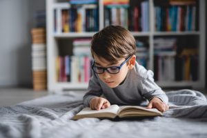 A child with brown hair wearing glasses reads a book while laying on the floor