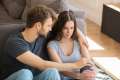 Man with his arm around a woman as they both sit on the ground in a living room