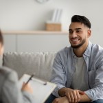 Happy middle eastern man talking to his psychologist, sharing therapy results with counselor, receiving professional help at mental health clinic