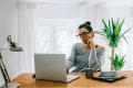 Woman with glasses resting her chin on her hand while looking at her laptop at a wooden desk in a bright home office