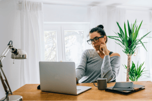 Woman with glasses resting her chin on her hand while looking at her laptop at a wooden desk in a bright home office