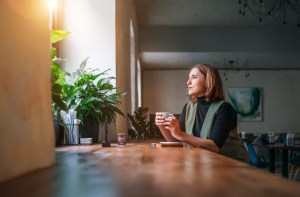 Woman sitting alone at a café table holding a coffee cup, gazing thoughtfully into the distance with her phone nearby