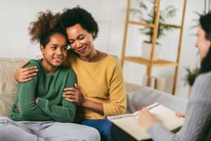 Woman in a yellow sweater and child in a green shirt on a gray couch, facing a therapist