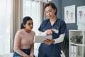 Woman sitting on an exam table talking to a nurse in navy blue scrubs holding a clipboard