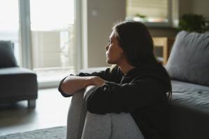 Stressed woman in gray sweatpants and a sweater sitting on the floor against a couch, staring out a window