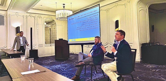 Two men in suits speaking in front of a white conference room with a projector behind them.