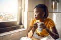 A young woman sitting by a sunlit window, holding a coffee mug and looking thoughtfully outside.