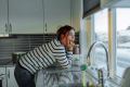 Woman in a striped sweater leaning on her kitchen counter, chin in hand, gazing out a window at a snowy view