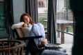 Red-haired woman sitting by a window drinking coffee and looking tired