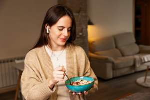 Woman in a tan sweater holding a blue ceramic bowl and eating granola with bluetooth earbuds in