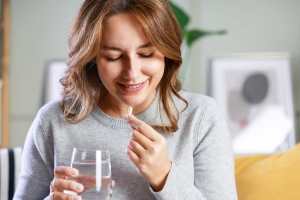 Woman in a gray sweater holding a glass of water taking a white pill