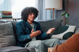 Young man sitting on couch at therapy session, arms out looking annoyed while sitting across from his therapist
