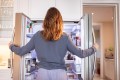 Woman standing in front of open fridge wearing pajamas, arms on both fridge doors. Inside is a packed fridge