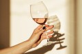 Close up of a woman's hand holding a glass of pink wine against a white wall