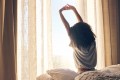 Woman sitting on bed, facing bright window, waking up and stretching