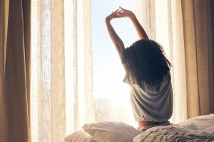 Woman sitting on bed, facing bright window, waking up and stretching